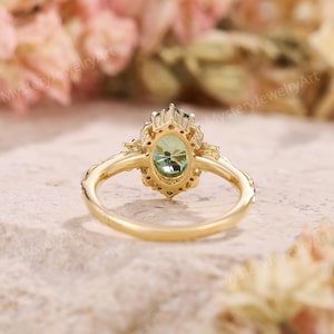 May include: A gold ring featuring an oval-cut light green gemstone surrounded by a halo of smaller stones. The band has a decorative, textured design on the sides. The text MysteryJewelryArt is visible in the background of the image.