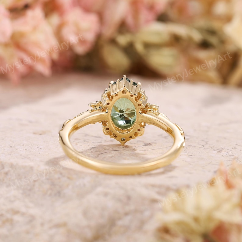 May include: A gold ring featuring an oval-cut light green gemstone surrounded by a halo of smaller stones. The band has a decorative, textured design on the sides. The text MysteryJewelryArt is visible in the background of the image.
