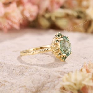 May include: A gold ring featuring a central oval-cut light green gemstone surrounded by smaller green stones. The band has a textured, vine-like design that extends into the setting, which holds the stones in a cluster arrangement.