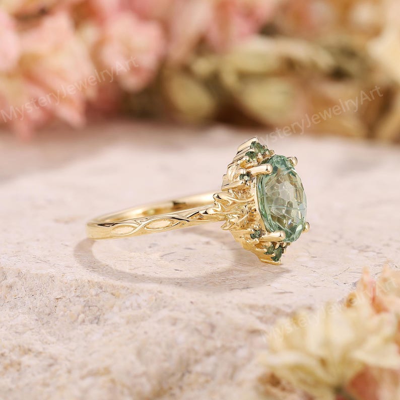 May include: A gold ring featuring a central oval-cut light green gemstone surrounded by smaller green stones. The band has a textured, vine-like design that extends into the setting, which holds the stones in a cluster arrangement.