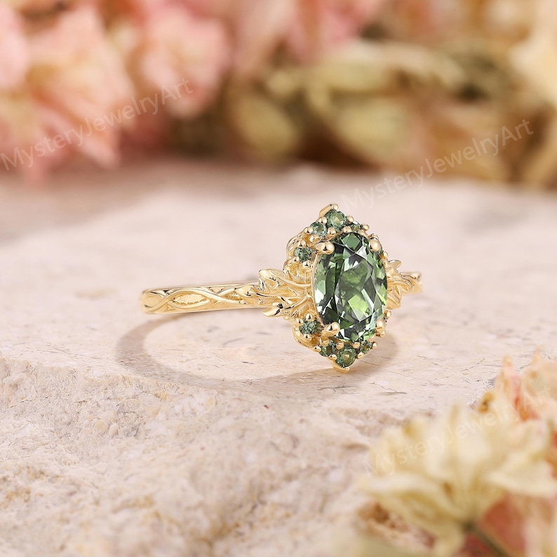 May include: A gold ring with a central oval green gemstone, surrounded by smaller green stones and ornate gold detailing. The band features a delicate, textured design. The ring is set against a soft, neutral background.