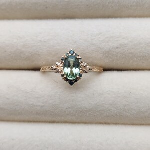 May include: A gold ring featuring an oval-cut, light green gemstone surrounded by smaller blue-green stones. The ring has an ornate, floral design on the band and around the central stone. The ring is set against a neutral background.
