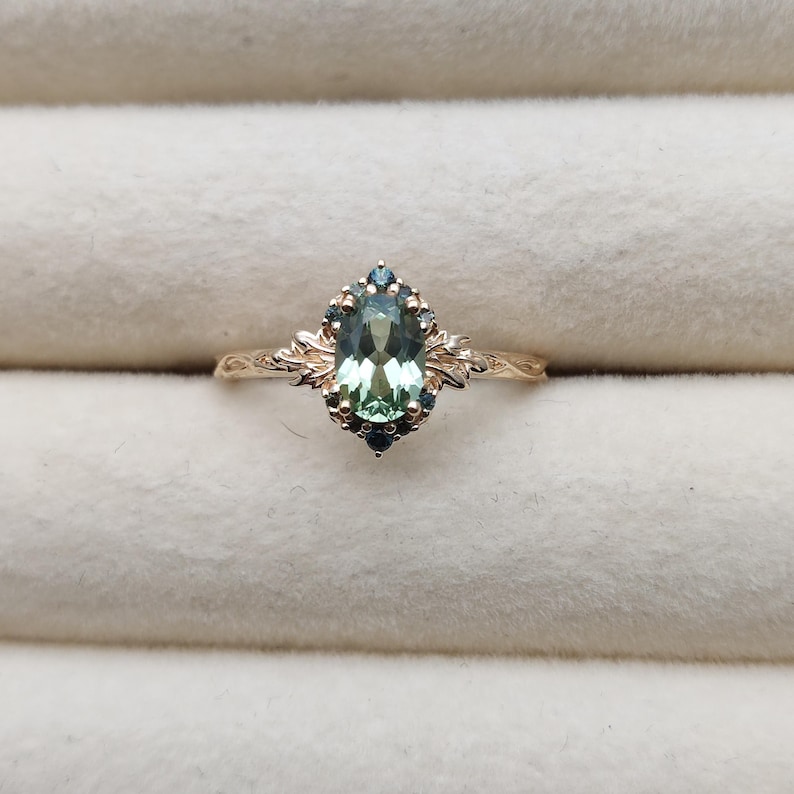 May include: A gold ring featuring an oval-cut, light green gemstone surrounded by smaller blue-green stones. The ring has an ornate, floral design on the band and around the central stone. The ring is set against a neutral background.