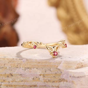 Peut inclure: Une bague dor&eacute;e avec un motif d&eacute;coratif tourbillonnant et trois petites pierres pr&eacute;cieuses rondes rouges. La bague est pos&eacute;e sur une surface textur&eacute;e de couleur claire.