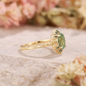 May include: A gold ring with an oval, light green gemstone at the center, surrounded by smaller green stones. The band has a delicate, engraved design. The ring is set against a neutral, textured background.