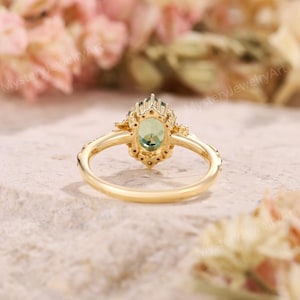 May include: A gold ring with an oval, light green gemstone at the center. The gemstone is surrounded by smaller, clear stones. The ring band is a simple, polished gold. The ring is set against a soft, blurred background.