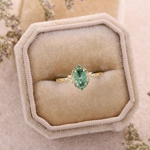 May include: A gold ring featuring a central oval-cut green gemstone surrounded by a halo of smaller green stones. The band is crafted with a detailed leaf-like texture, and the setting includes delicate metal prongs securing the stones in place.