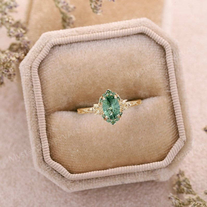 May include: A gold ring featuring a central oval-cut green gemstone surrounded by a halo of smaller green stones. The band is crafted with a detailed leaf-like texture, and the setting includes delicate metal prongs securing the stones in place.