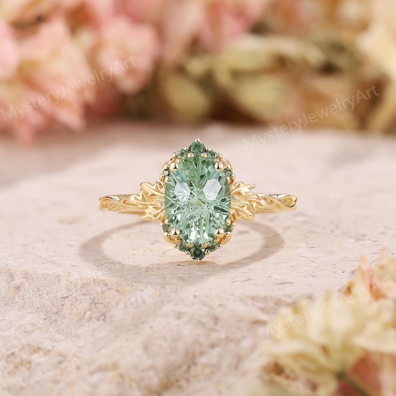 May include: A gold ring with a light green oval-cut gemstone at the center, surrounded by smaller green stones. The ring has a detailed floral design on the band and around the central stone. The ring is set on a textured, light-colored surface.