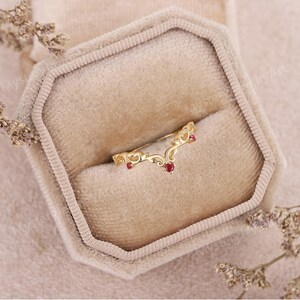 Peut inclure: Une bague dor&eacute;e en forme de V avec des d&eacute;tails de filigrane orn&eacute;s. La bague comporte trois petites pierres pr&eacute;cieuses rondes rouges. La bague est pr&eacute;sent&eacute;e dans un &eacute;crin en velours beige.