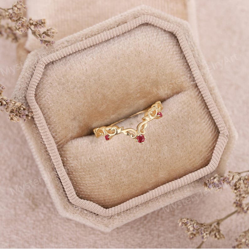 Peut inclure: Une bague dor&eacute;e en forme de V avec des d&eacute;tails de filigrane orn&eacute;s. La bague comporte trois petites pierres pr&eacute;cieuses rondes rouges. La bague est pr&eacute;sent&eacute;e dans un &eacute;crin en velours beige.