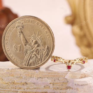 Peut inclure: Une bague dor&eacute;e avec un design incurv&eacute; et trois petites pierres pr&eacute;cieuses rouges. La bague est &agrave; c&ocirc;t&eacute; d'une pi&egrave;ce d'un dollar am&eacute;ricain, montrant la Statue de la Libert&eacute;. La bague est sur une surface textur&eacute;e et claire.