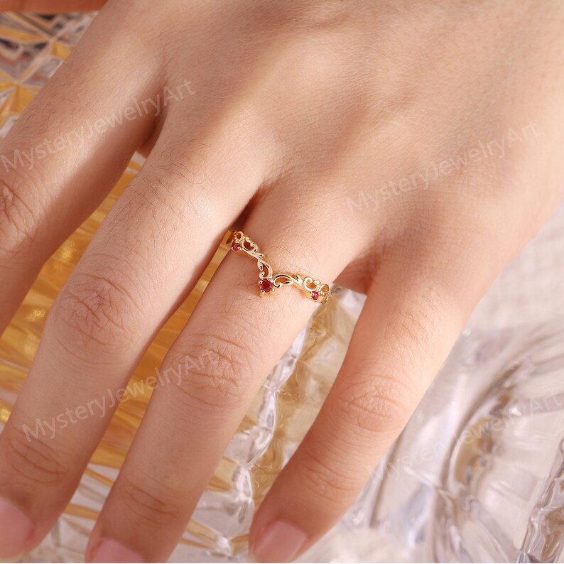 Peut inclure: Une bague dor&eacute;e avec un design ajour&eacute; d&eacute;licat. La bague pr&eacute;sente une forme en V orn&eacute;e de petites pierres rouges. La bague est port&eacute;e sur un doigt, mettant en valeur les d&eacute;tails et le savoir-faire de la joaillerie.