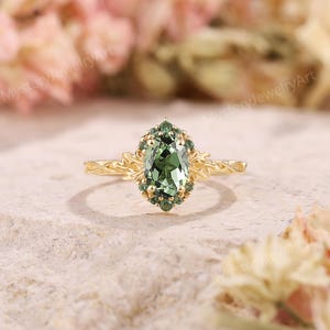 May include: A gold ring with an oval, green gemstone center, surrounded by smaller green stones. The ring's band features an intricate, floral design. The ring is set against a soft, neutral background, highlighting its elegant details.