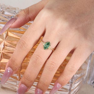 May include: A gold ring featuring a marquise-cut green gemstone set in a textured band. The ring is worn on a finger, showcasing the intricate metalwork surrounding the central stone and the detailed, branch-like design of the band itself.