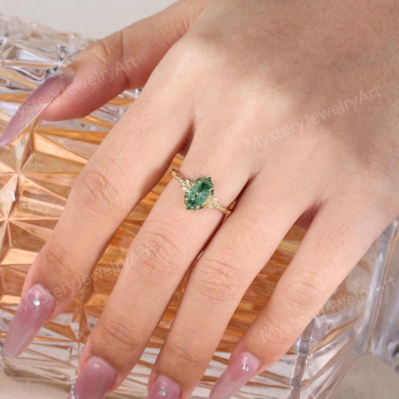 May include: A gold ring featuring a marquise-cut green gemstone set in a textured band. The ring is worn on a finger, showcasing the intricate metalwork surrounding the central stone and the detailed, branch-like design of the band itself.