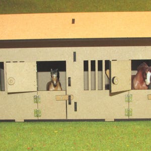 May include: A light brown wooden toy horse stable with a slanted roof, featuring two stalls with hinged doors and vertical slats. Inside each stall, a small plastic horse figurine is visible. The structure is constructed from flat panels.