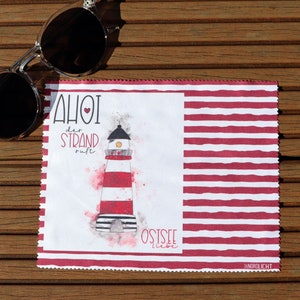 Op de afbeelding: Een witte microvezel schoonmaakdoek met rode strepen. De doek heeft een illustratie van een vuurtoren met de tekst "AHOI der STRAND ruft OSTSEE liebe" in rood.