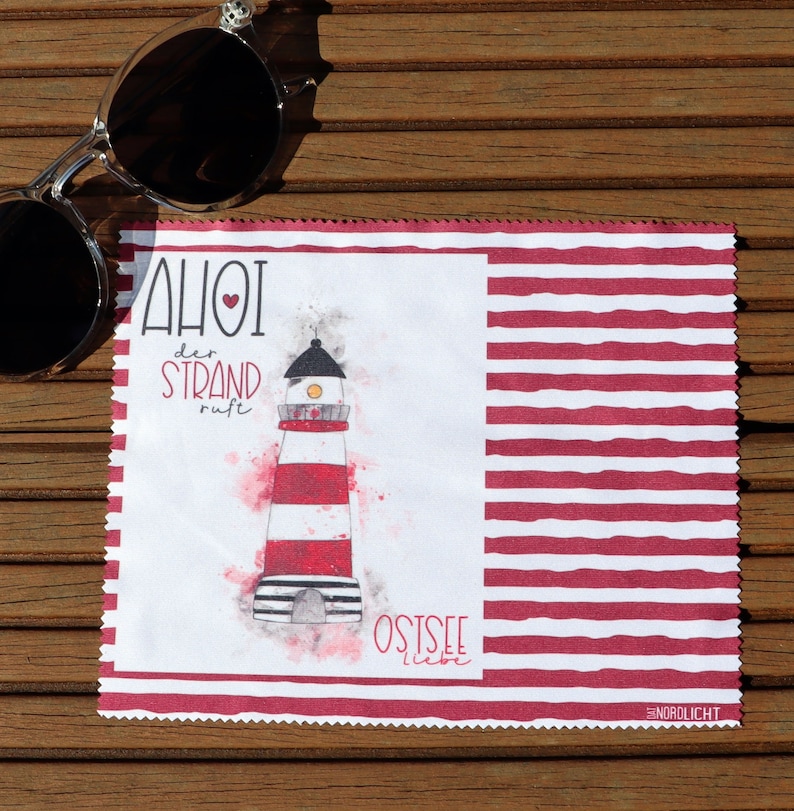 Op de afbeelding: Een witte microvezel schoonmaakdoek met rode strepen. De doek heeft een illustratie van een vuurtoren met de tekst "AHOI der STRAND ruft OSTSEE liebe" in rood.