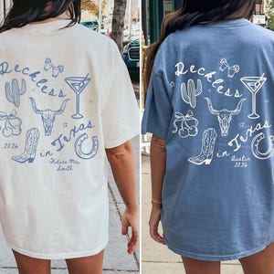 May include: An ivory t-shirt and a blue t-shirt, each featuring a back graphic with the text "Reckless in Texas". The design includes illustrations of a cactus, a martini glass, a cow skull, a pair of cherries, a cowboy boot, and a horseshoe.
