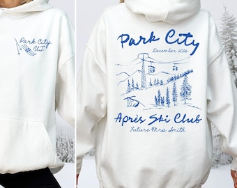 May include: A white hooded sweatshirt featuring blue ink graphics. The front displays 'Park City, UT' with ski icons. The back shows a mountain landscape with a ski lift, pine trees, and the text 'Park City', 'December 2026', 'Après Ski Club', and 'Future Mrs. Smith'.