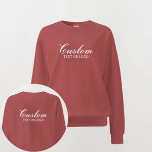 May include: A brick red crewneck sweatshirt featuring the white text Custom TEXT OR LOGO printed on the chest. The garment has long sleeves and a ribbed neckline, cuffs, and hem. A circular inset shows the same sweatshirt design in a smaller view.