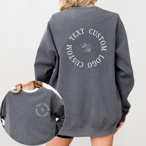 May include: A charcoal gray crewneck sweatshirt featuring a circular white graphic on the back and a smaller matching version on the front left chest. The design includes the text CUSTOM TEXT CUSTOM LOGO arranged in a circle with script initials.