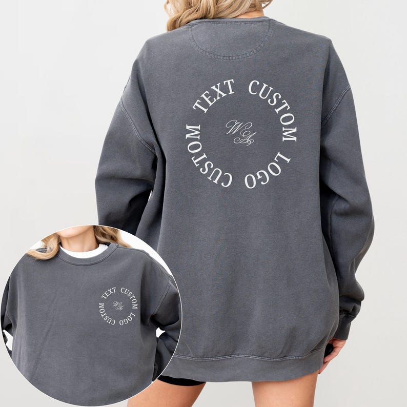May include: A charcoal gray crewneck sweatshirt featuring a circular white graphic on the back and a smaller matching version on the front left chest. The design includes the text CUSTOM TEXT CUSTOM LOGO arranged in a circle with script initials.