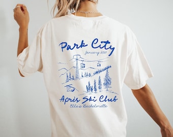 Puede incluir: Una camiseta color hueso con un gráfico de líneas azules en la espalda. El diseño muestra un paisaje de montaña con telesillas, pinos y el texto Park City, January 2021, Après Ski Club y Ella's Bachelorette.