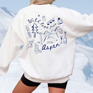 May include: A white long-sleeve sweatshirt featuring a dark blue line art graphic on the back. The design includes illustrations of skis, a pine tree, a ski lift, a beanie, mountains, a ski boot, and a champagne bottle with the text With love from Aspen 2026.