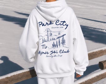 Puede incluir: Una sudadera con capucha blanca que presenta un gráfico azul oscuro en la parte posterior. El diseño incluye el texto Park City, December 2026, y Après Ski Club, Family Ski Trip, junto a una ilustración de montañas, telesillas y pinos.