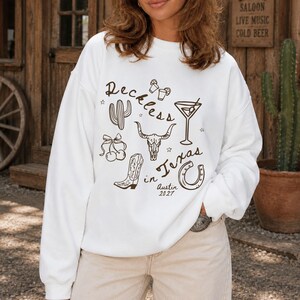 May include: A white long-sleeve sweatshirt featuring a brown graphic print with the text "Reckless in Texas" and "Austin 2027". The design includes illustrations of a cactus, a martini glass, a cow skull, a cowboy boot, a horseshoe, and cherries.
