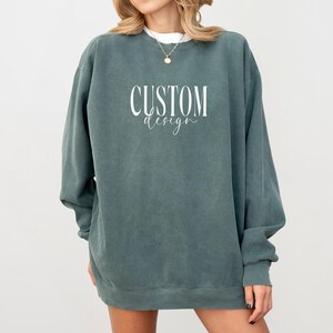 May include: A dark teal crewneck sweatshirt featuring the white text "CUSTOM design" printed on the chest. The garment has long sleeves, a ribbed neckline, and a relaxed fit. The fabric has a slightly faded, vintage-inspired appearance.