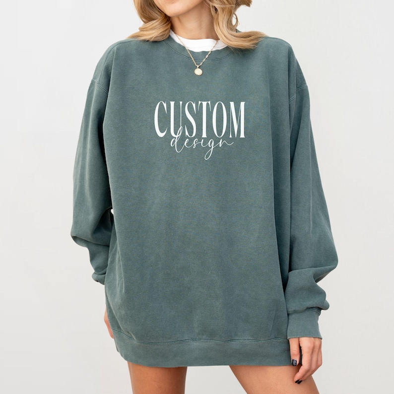 May include: A dark teal crewneck sweatshirt featuring the white text "CUSTOM design" printed on the chest. The garment has long sleeves, a ribbed neckline, and a relaxed fit. The fabric has a slightly faded, vintage-inspired appearance.