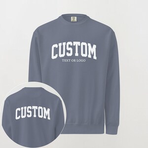 May include: A slate blue crewneck sweatshirt featuring white collegiate-style lettering that reads CUSTOM TEXT OR LOGO on the chest. A circular inset shows the back of the same sweatshirt with the word CUSTOM printed in white across the upper back.