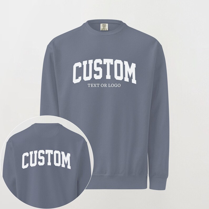 May include: A slate blue crewneck sweatshirt featuring white collegiate-style lettering that reads CUSTOM TEXT OR LOGO on the chest. A circular inset shows the back of the same sweatshirt with the word CUSTOM printed in white across the upper back.