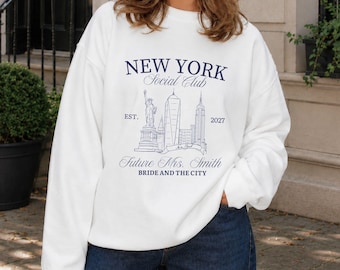 May include: A white crewneck sweatshirt featuring a dark blue graphic print of the New York City skyline, including the Statue of Liberty. The text reads NEW YORK Social Club, EST. 2027, Future Mrs. Smith, and BRIDE AND THE CITY in a serif font.