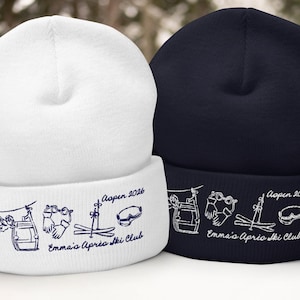 May include: Two beanies, one white and one navy blue, are displayed. Both have a folded brim with a ski-themed design and the text "Aspen 2026" and "Emma's Aprio Ski Club". The design includes ski lift, goggles, skis, and gloves.