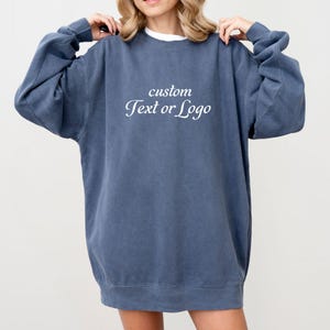 May include: A slate blue crewneck sweatshirt featuring a relaxed fit with long sleeves and a ribbed hem. The center of the chest displays the white text "custom Text or Logo" in a serif font. The garment has a washed, vintage-inspired appearance.