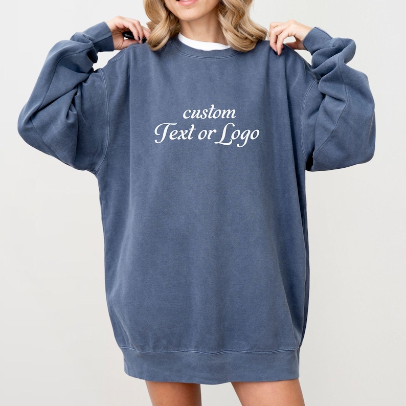 May include: A slate blue crewneck sweatshirt featuring a relaxed fit with long sleeves and a ribbed hem. The center of the chest displays the white text "custom Text or Logo" in a serif font. The garment has a washed, vintage-inspired appearance.