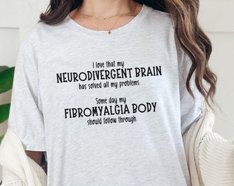 Puede incluir: Una camiseta de color gris jaspeado claro con texto en negro que dice: I love that my NEURODIVERGENT BRAIN has solved all my problems. Some day my FIBROMYALGIA BODY should follow through. La prenda tiene mangas cortas y cuello redondo.