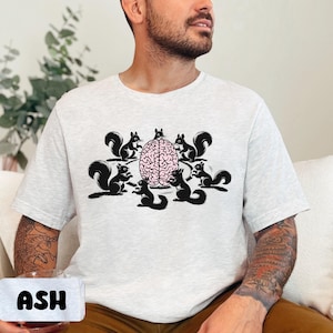 Op de afbeelding: Een lichtgrijs T-shirt met een afbeelding van vijf eekhoorns rond een roze brein. De eekhoorns zijn zwart en wit.