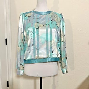 Peut inclure: Un chemisier vintage en satin dans les tons de bleu, turquoise et blanc, présenté sur un mannequin. Le haut à manches longues présente un motif à rayures avec des rayures verticales turquoise et des motifs abstraits au pinceau. Le chemisier a un col rond et des poignets et un ourlet élastiqués.