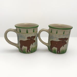 May include: Two ceramic mugs with a nature theme. Each mug features a brown moose illustration on a light background with green grass and trees. The rims are green, and the handles are beige.