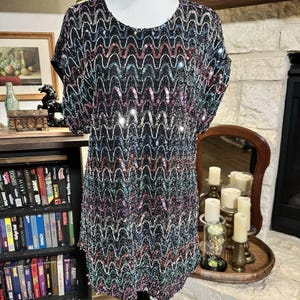 Puede incluir: Vestido negro con lentejuelas con un patrón de ondas en tonos rosa, azul y plateado. El vestido tiene mangas cortas y cuello redondo. El vestido se exhibe en un maniquí.
