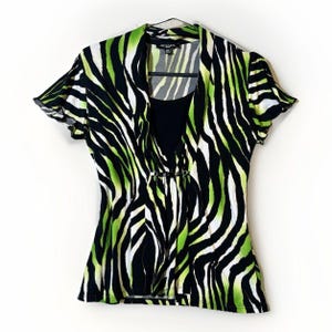 May include: A short-sleeved top with a zebra print in black, white, and lime green. The top has a layered design with a black inner layer and a draped outer layer. The outer layer is secured with a decorative clasp.