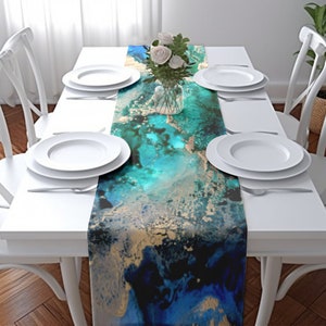 May include: A rectangular table runner with an abstract design in shades of teal, gold, and navy blue. The runner is placed on a white table set with white plates, silverware, and a vase of white roses. The table is surrounded by white chairs.