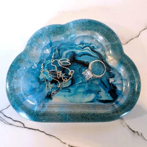 May include: A cloud-shaped trinket tray with a blue and white marbled design. The tray holds a pair of silver bunny earrings and a ring with a large, square gemstone. The tray's edge is lined with glitter.