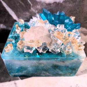 May include: A teal blue resin box with a clear lid. The lid is decorated with a cluster of clear and gold glitter, a large clear quartz crystal, and a cluster of teal blue crystals.