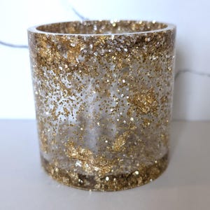 May include: A cylindrical, clear resin container with gold glitter and gold leaf accents. The container is a decorative item, possibly a candle holder or pen holder. The gold elements are distributed throughout the resin, creating a sparkling effect.